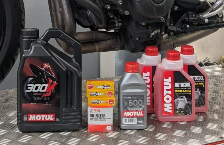 Various bottles of engine oil and a box of oil filters lined up next to a motorcycle engine.