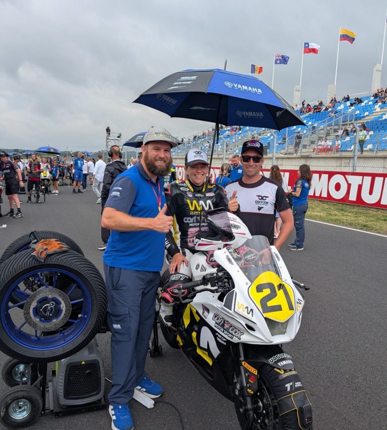 Three people (Avalon Lewis, Jake Lewis and Scoot) pose with a motorcycle under an umbrella at a racing event.