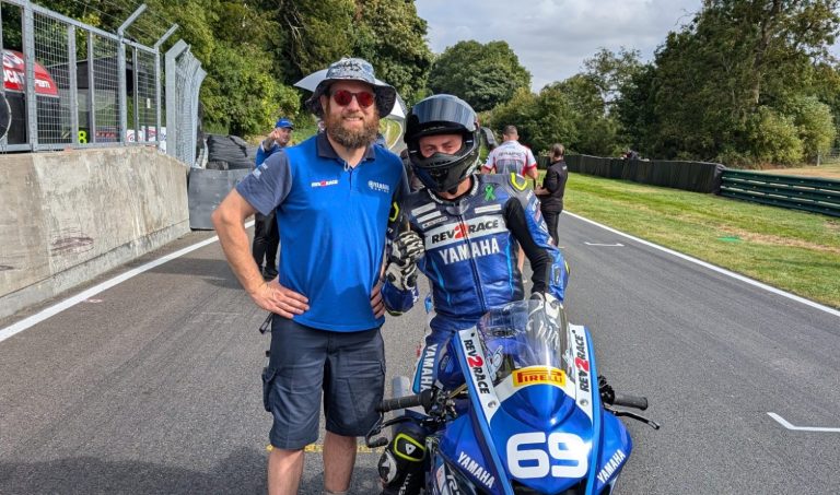 Two men pose on a racetrack, one in motorcycle gear and the other in casual attire.