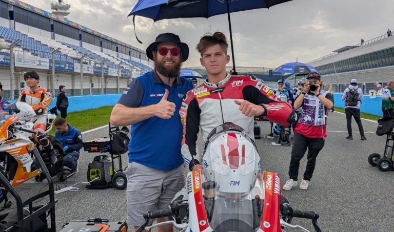 Two people (Blake Davis and Scoot), one in racing gear and the other in casual attire, pose with a motorcycle.